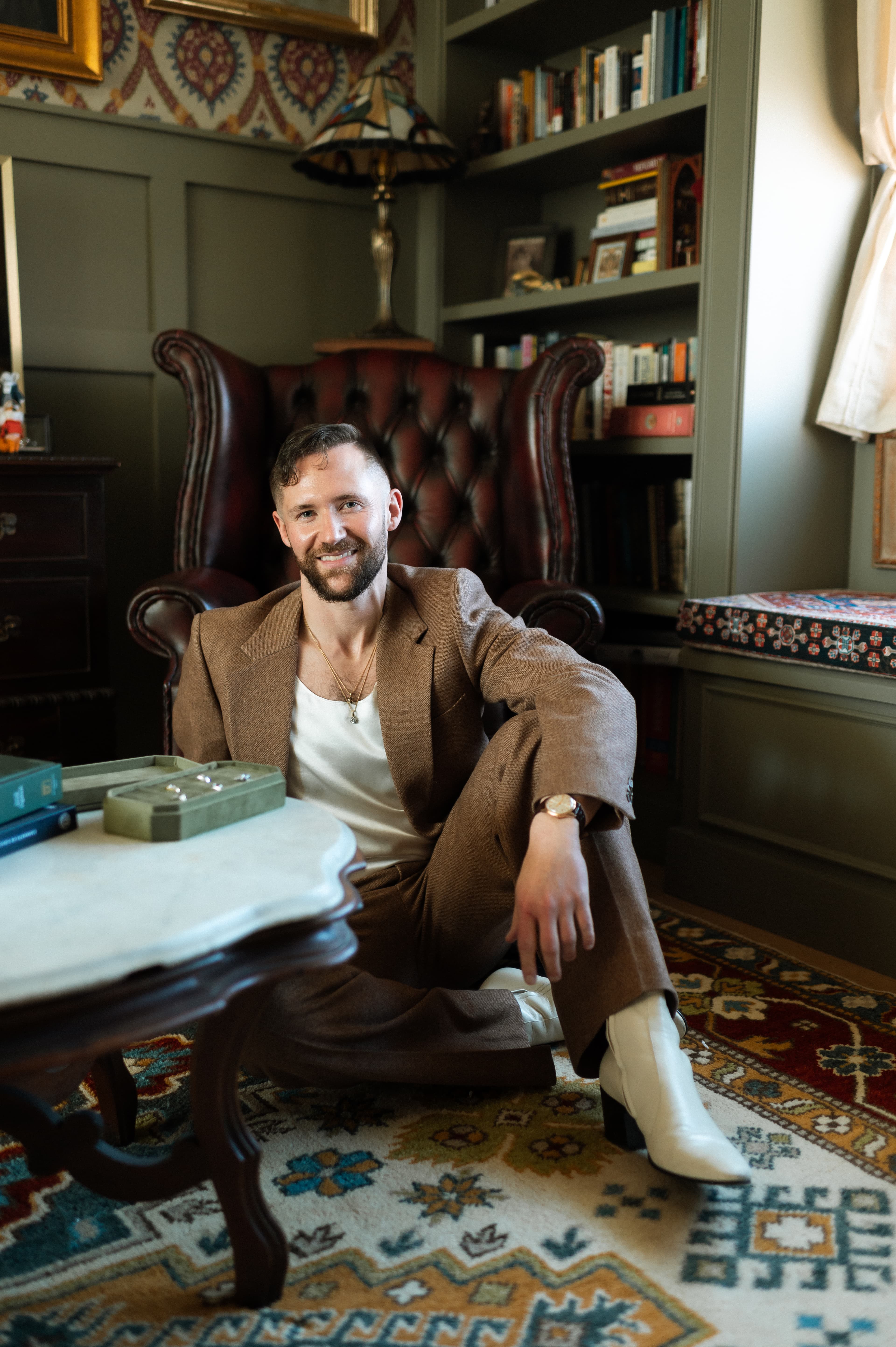 Marc Baker-Bourgeois seated in a leather wingback chair in his Denver atelier, wearing a tailored brown suit